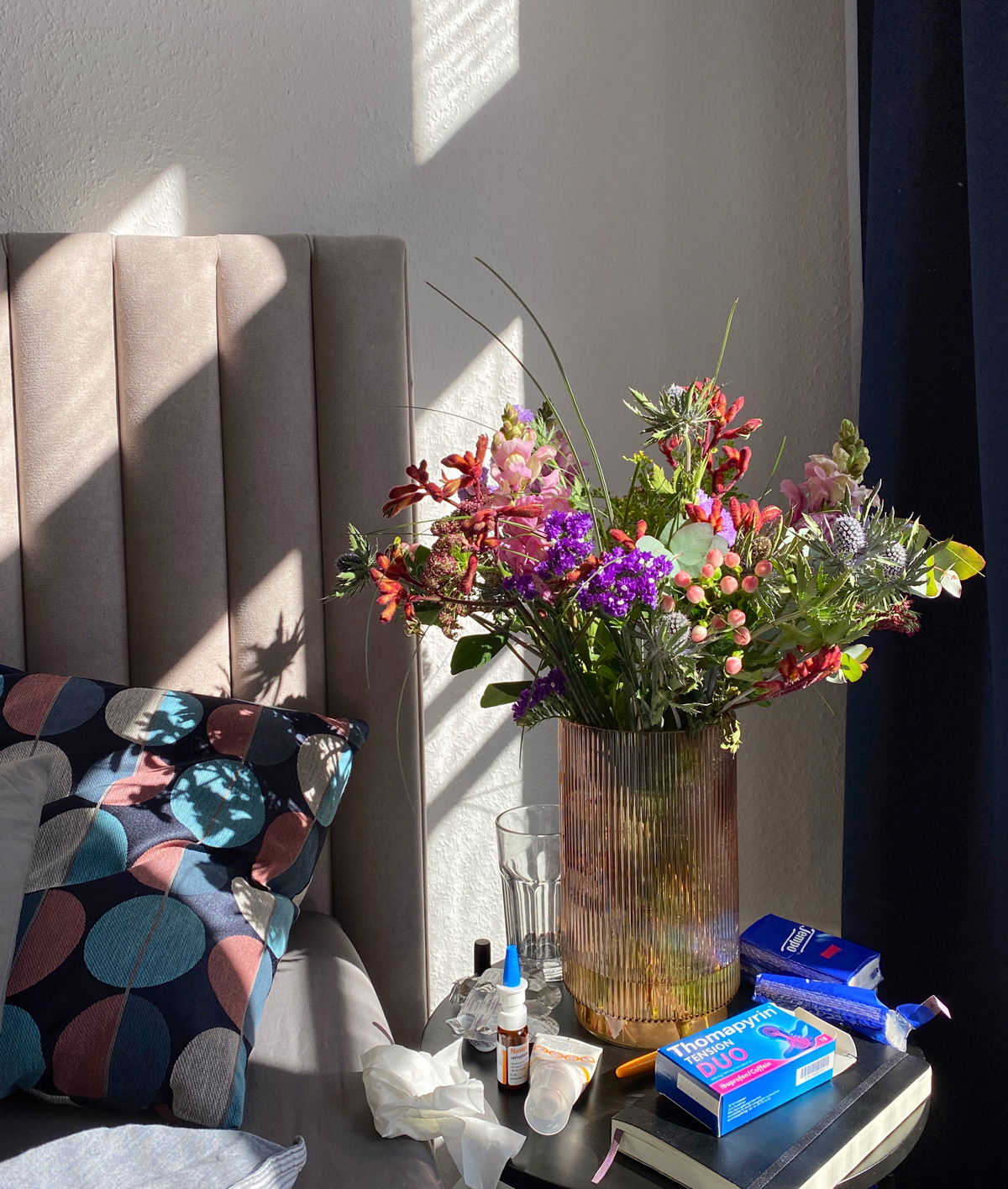 Foto von einem großen Blumenstrauß auf dem Nachtisch eines Bettes. Daneben liegen Taschentücher und Schmerztabletten